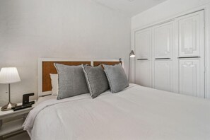 Cama bem arrumada com cabeceira, luminária de cabeceira e mesa de apoio com telefone e controle remoto.