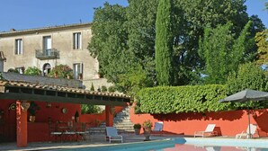 Une piscine avec des chaises longues, un bâtiment rouge et un grand arbre en arrière-plan.