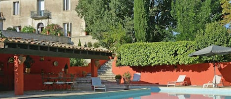 A swimming pool with lounge chairs, a red building, and a tall tree in the background.
