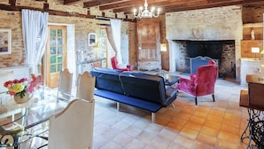 A rustic interior with a stone fireplace, wooden beams, a sofa, and a dining area with a glass table and chairs.