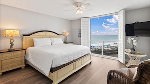 A bedroom with a large bed, a ceiling fan, a nightstand with a lamp, a wicker chair, and a view of the beach through an open window. - Beautiful OCEAN VIEW! 2 FREE Beach chairs and umbrella! (Destin)