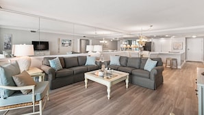 A modern living room with a sectional sofa, a glass coffee table, and a dining area with a kitchen in the background. - Beautiful OCEAN VIEW! 2 FREE Beach chairs and umbrella! (Destin)