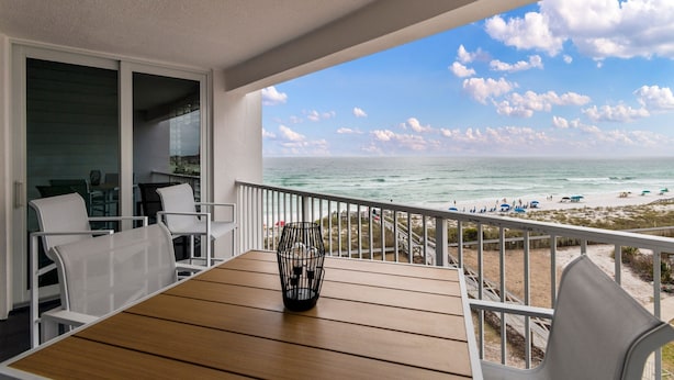 A balcony with a wooden table and chairs, overlooking a beach with people and umbrellas. - Beautiful OCEAN VIEW! 2 FREE Beach chairs and umbrella! (Destin)