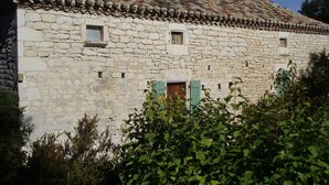 Un edificio de piedra con techo de tejas, dos ventanas pequeñas y una puerta de madera con contraventanas verdes.