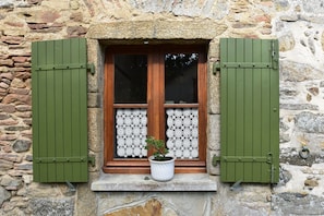 Un edificio de piedra con una ventana que tiene contraventanas verdes, una planta en maceta y cortinas de encaje.
