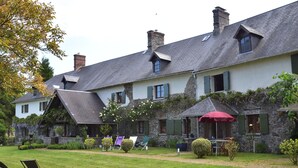 A stone building with a sloped roof, multiple chimneys, and a garden with outdoor seating. - Saint-Sauveur Hideaway (St Sauveur Lendelin)