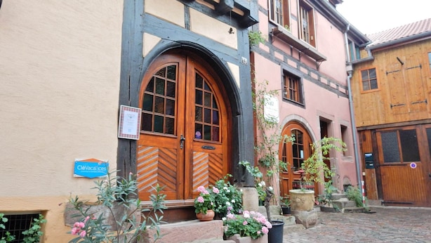 Un bâtiment traditionnel doté d’une grande porte en bois et de fenêtres en arc de cercle, entouré de plantes en pot et bordé d’une rue pavée.