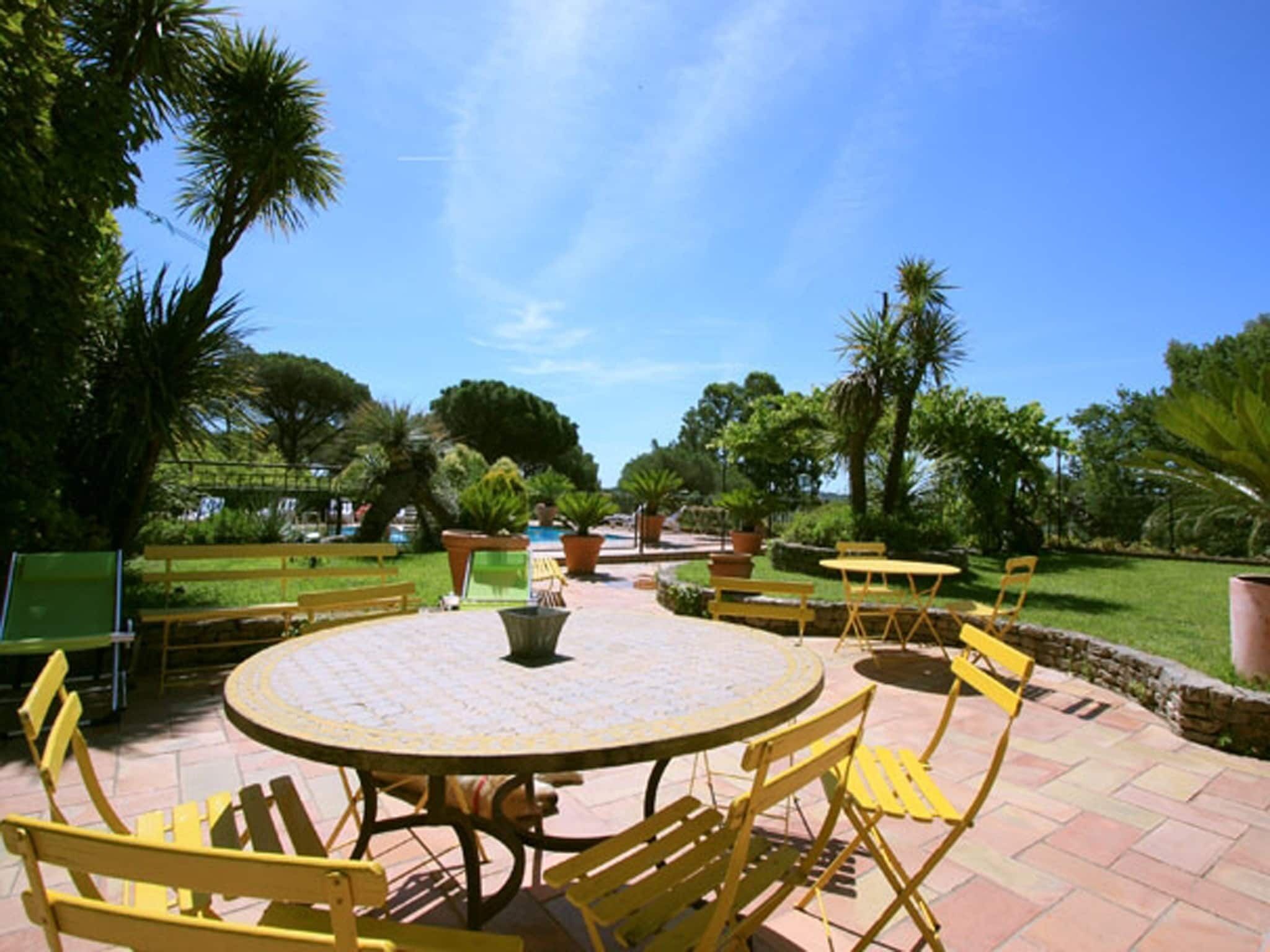 An outdoor patio with yellow chairs and a round table, surrounded by greenery and palm trees.