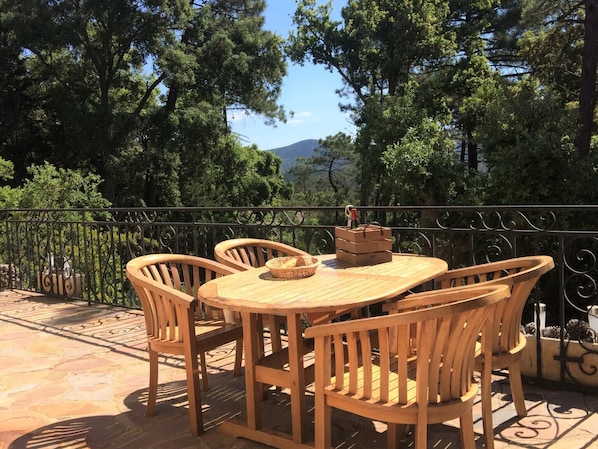 An outdoor wooden table and chairs set on a terrace with a metal railing, surrounded by trees and a mountainous backdrop.
