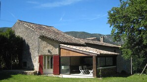 Une maison en pierre avec un toit en tuiles, une porte en verre et un petit espace extérieur aménagé avec des chaises.
