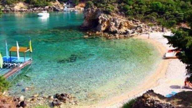 Une plage côtière aux eaux turquoise et limpides, bordée d’une côte rocheuse et dotée d’un petit quai où est amarré un bateau.