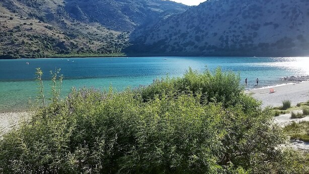 A mountainous landscape with a clear lake, a beach, and some vegetation in the foreground.