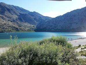 A mountainous landscape with a clear lake, a beach, and some vegetation in the foreground.