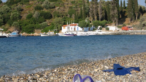 A pebbled beach with boats docked in the background and a building with a red roof. - Villa in Samos near Aghia Paraskevi Beach (Aghia Paraskevi)