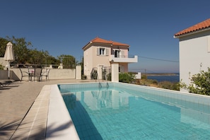 A swimming pool with a clear view of the sea, a two-story building with a red-tiled roof, and outdoor furniture.