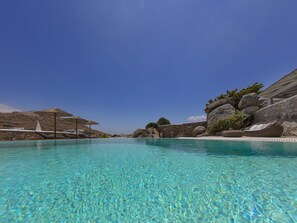 Ein klares, blaues Schwimmbecken mit Liegestühlen und Sonnenschirmen, umgeben von felsigem Gelände und Steinmauern.