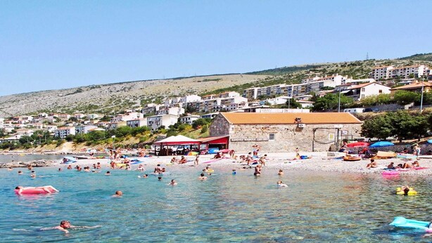 A beach with clear water, people swimming, and buildings in the background. - Apartment in Senj with Sea Views & BBQ (Senj)