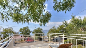 A balcony with two wooden lounge chairs, a tiled floor, and a view of parked cars and trees. - Apartment in Senj with Sea Views & BBQ (Senj)