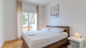 A neatly made bed with white linens in a room with wooden flooring, a bedside table with a lamp, and a window with curtains. - Apartment in Senj with Sea Views & BBQ (Senj)