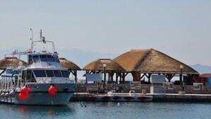 Un bateau était amarré à un quai, bordé de constructions aux toits de chaume, avec une chaîne de montagnes en arrière-plan.
