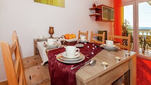 Un comedor con una mesa dispuesta para cuatro personas, una mesita auxiliar con una cesta de naranjas y un cuadro en la pared.