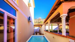 Ein schmaler Pool mit Fliesenveranda, umgeben von einem Gebäude mit einem gefliesten Dach und weißen Säulen.
