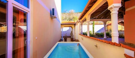A narrow swimming pool with a tiled deck, surrounded by a building with a tiled roof and white columns.
