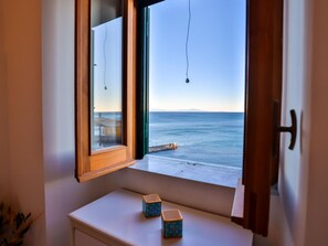 A room with a view of the sea through an open window, a small table with two blue containers, and a wooden door.