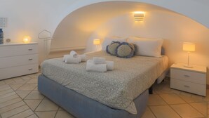 Un dormitorio moderno con techo curvo, una cama grande con ropa de cama estampada, dos lámparas de noche y una mesita de noche con lámpara.