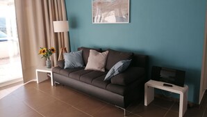 A living room with a brown sofa, a small white side table, a lamp, a framed picture on the wall, and a TV on a stand. - Apartment by the Beach in Dalmatia (Šibenik)