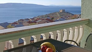 Un balcon avec une table, une chaise et une vue sur une ville côtière, avec ses maisons et son château.