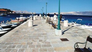 Une promenade en bord de mer, dallée de pierres, agrémentée de bancs et de lampadaires, avec des bateaux amarrés le long du quai, et offrant une vue dégagée sur la mer et les montagnes lointaines.