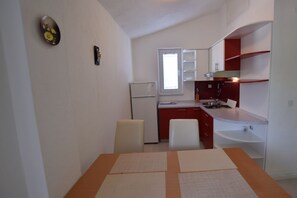 Een kleine keuken met witte kasten, een koelkast en een raam. Er staat een eettafel met stoelen.