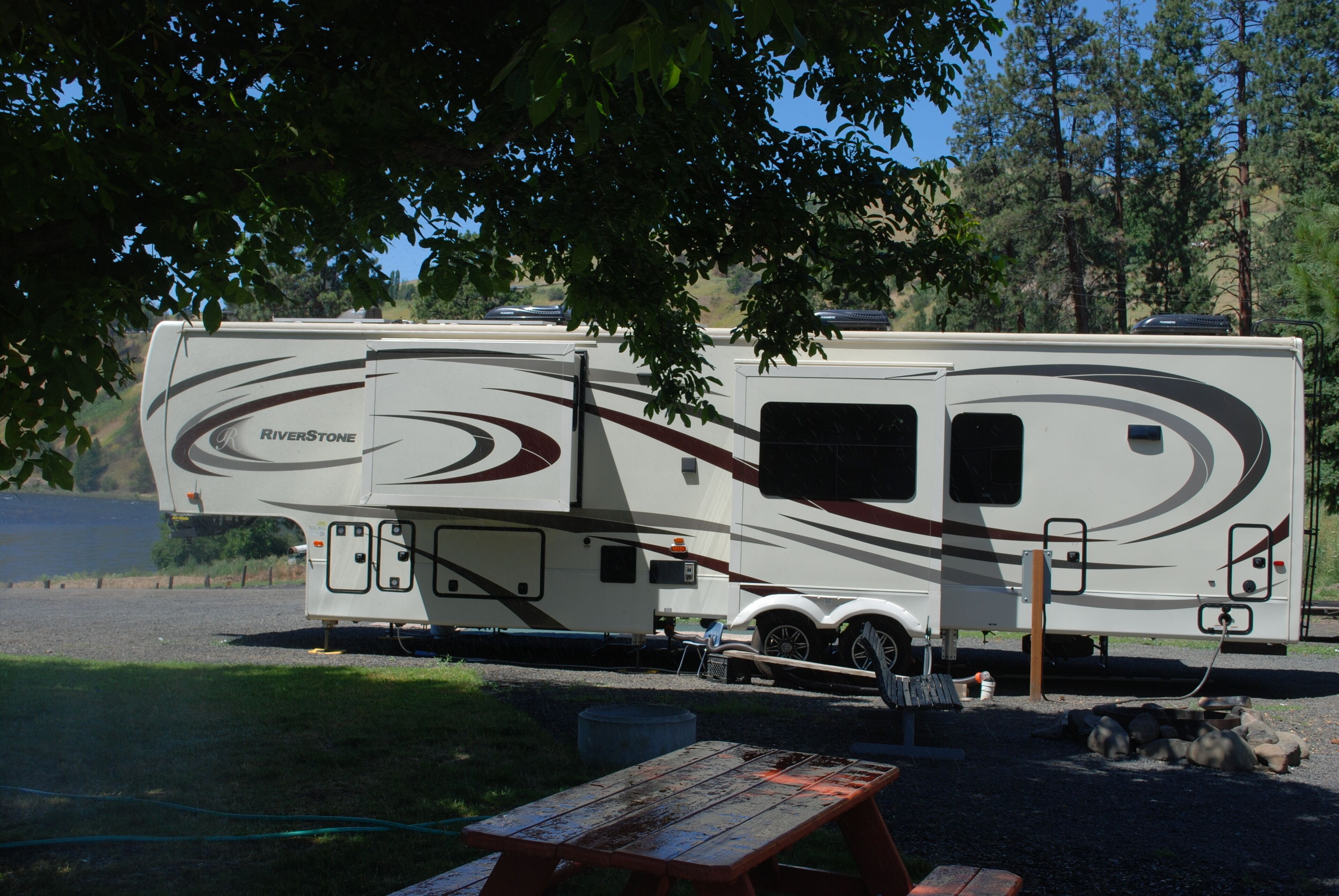 A RiverStone RV parked by a lake with picnic tables nearby.