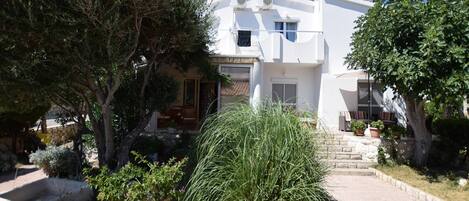 A white building with a balcony, surrounded by trees and potted plants.