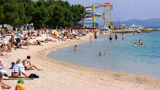 Une plage bondée où les gens se prélassent au soleil et se baignent, avec un toboggan aquatique et des bateaux en arrière-plan.