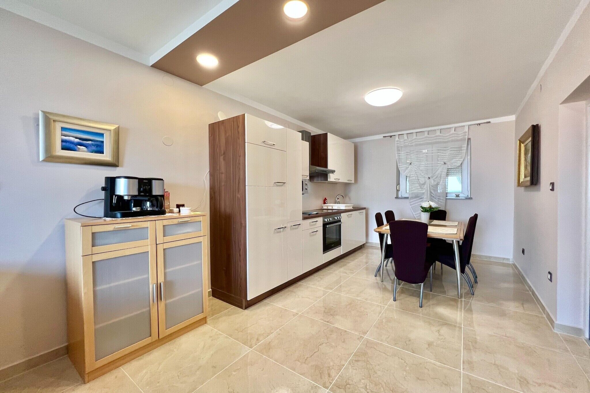 A modern kitchen with a dining area, featuring a coffee maker, cabinets, and a window with curtains.