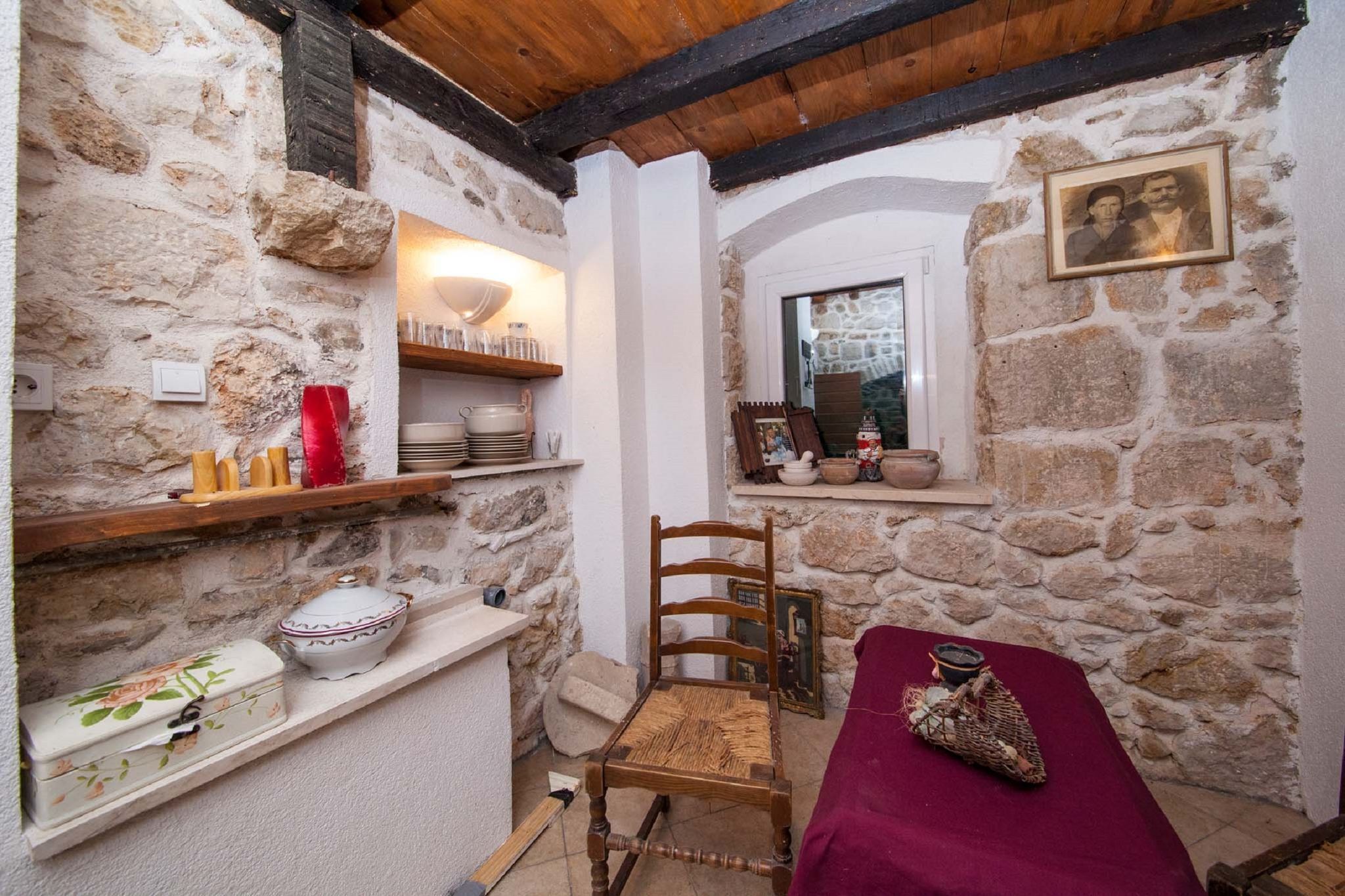 A rustic room with stone walls, a wooden chair, a bed with a red blanket, and shelves with kitchenware.