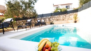 Un espace aménagé au bord de la piscine, avec un grand saladier en verre rempli de fruits, notamment des bananes, des pommes et des raisins, et un mur de pierre surmonté d’un toit en tuiles en arrière-plan.