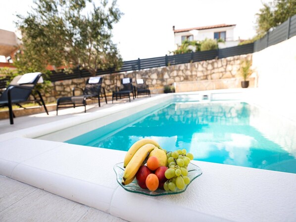 A poolside area with a glass bowl of fruit, including bananas, apples, and grapes, and a stone wall with a tiled roof in the background.