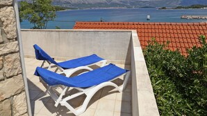 A balcony with two white lounge chairs covered with blue towels, overlooking a coastal area with a red-tiled roof and a view of the sea and mountains. - Holiday Home in Lumbarda with Garden (Lumbarda)