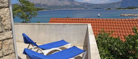 A balcony with two white lounge chairs covered with blue towels, overlooking a coastal area with a red-tiled roof and a view of the sea and mountains.