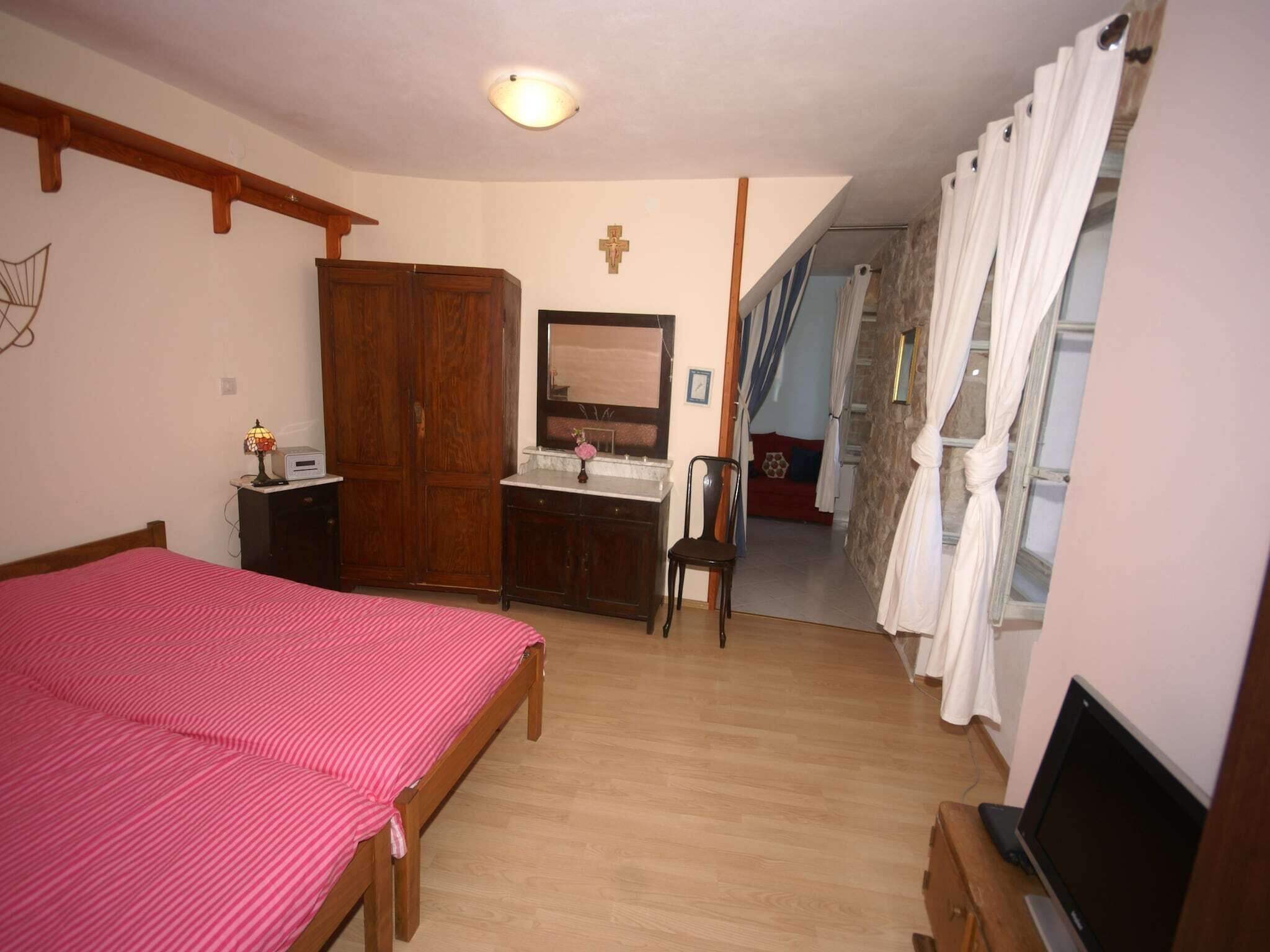 A bedroom with a bed, a wooden wardrobe, a desk with a mirror, a chair, and a television.