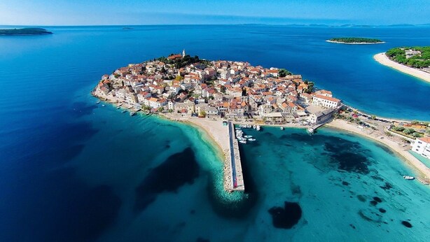 Vue aérienne d’une ville côtière avec une île centrale, entourée d’eaux d’un bleu limpide et d’une jetée qui s’avance dans la mer.