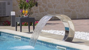 Une piscine agrémentée d’un élément décoratif aquatique, un mur en pierre et une table ornée de fleurs et de fruits.