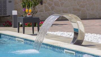 Une piscine agrémentée d’un élément décoratif aquatique, un mur en pierre et une table ornée de fleurs et de fruits.