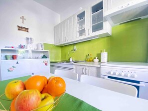 A kitchen with white cabinets, a green backsplash, and a sink. There is a fruit bowl with apples and oranges on the counter.