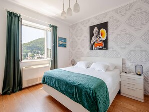 A bedroom with a large bed, a window with curtains, a radiator, a bedside table with a lamp, and a wall with patterned wallpaper and artwork.
