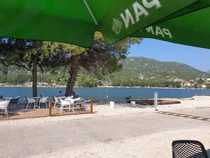 A seaside outdoor seating area with tables and chairs under a green umbrella, overlooking a body of water and a hillside.