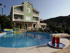 A swimming pool with a lounge area, a multi-story building with balconies, and a mountainous backdrop.